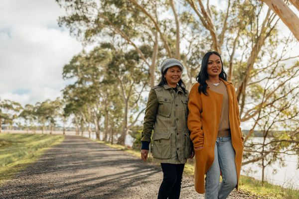 Two ladies walk on a gravel path, dressed in winter clothes. On their right is a row of trees separating them from the edge of the reservoir.
